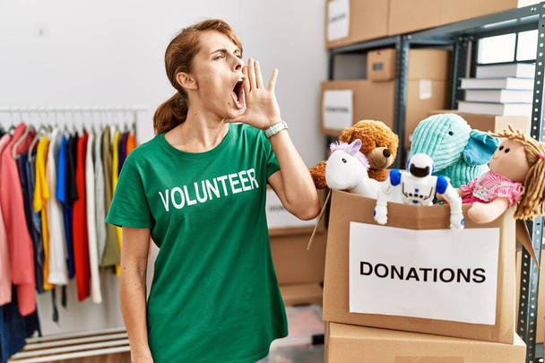 Красивая белая женщина в футболке добровольца на пожертвования стоять кричать и громко кричать бок о бок с рукой в рот. коммуникационная концепция.  - Фото, изображение