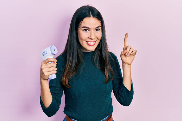 Young brunette woman holding thermometer smiling with an idea or question pointing finger with happy face, number one  - Photo, Image