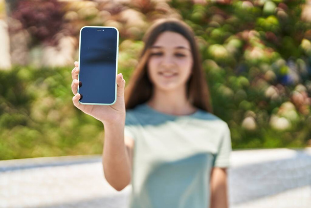 Schattig meisje glimlachend zelfverzekerd tonen scherm smartphone in het park - Foto, afbeelding