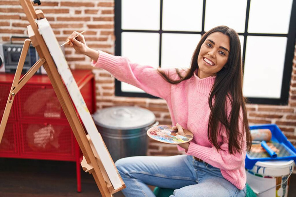 Genç İspanyol kadın resim stüdyosunda kendine güvenen gülümsüyor. - Fotoğraf, Görsel