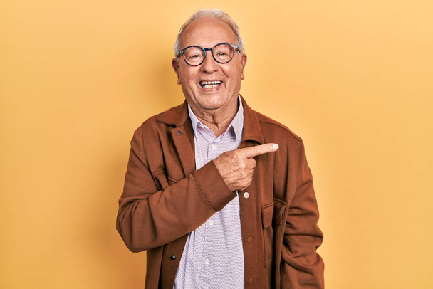 Älterer Mann mit grauen Haaren trägt lässige Jacke und Brille fröhlich mit einem Lächeln im Gesicht, das mit der Hand und dem Finger zur Seite zeigt, mit glücklichem und natürlichem Gesichtsausdruck  - Foto, Bild