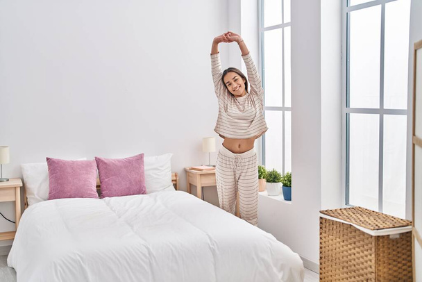 Young hispanic woman stretching arms standing at bedroom - Fotó, kép