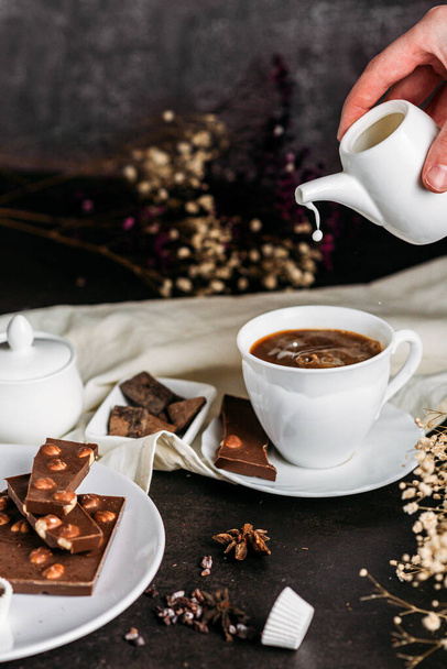 Kaffee in einer weißen Tasse mit Schokoladenblüten Hand gießt Milch aus Milchkraut Gefriertropfen schwarzen Hintergrund - Foto, Bild