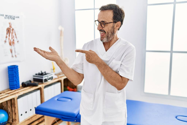 Middle age man with beard working at pain recovery clinic amazed and smiling to the camera while presenting with hand and pointing with finger.  - Foto, Imagen