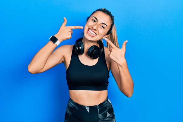 Beautiful hispanic woman wearing gym clothes and using headphones smiling cheerful showing and pointing with fingers teeth and mouth. dental health concept.  - Fotografie, Obrázek