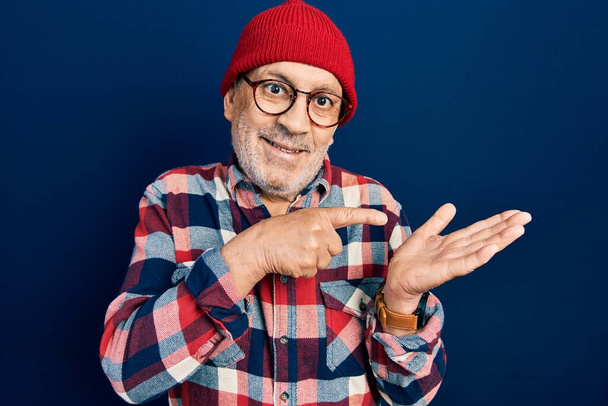Handsome mature man wearing hipster look with wool cap amazed and smiling to the camera while presenting with hand and pointing with finger.  - 写真・画像