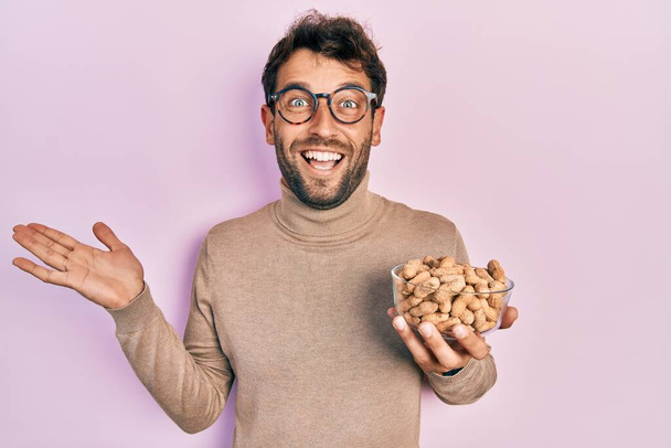 Handsome man with beard holding peanuts celebrating achievement with happy smile and winner expression with raised hand  - Photo, Image