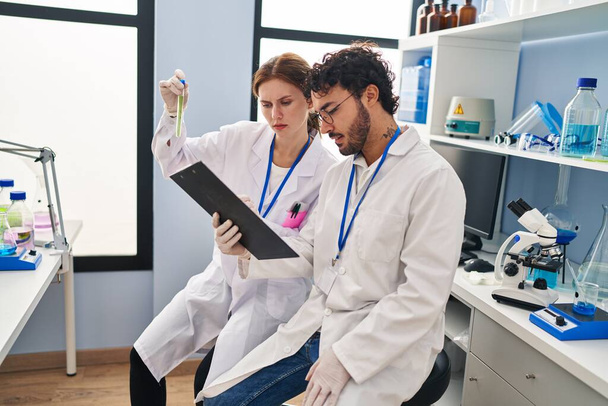Homem e mulher cientistas parceiros olhando tubo de ensaio segurando prancheta no laboratório - Foto, Imagem