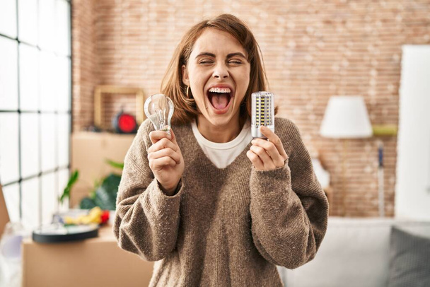 Young beautiful woman holding led lightbulb and incandescent bulb smiling and laughing hard out loud because funny crazy joke.  - Foto, immagini