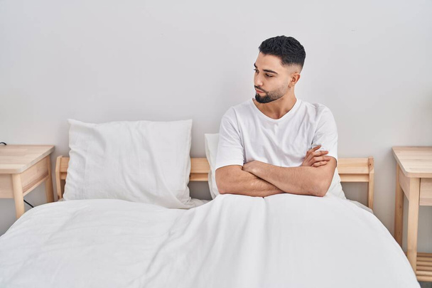 Hombre árabe joven sentado en la cama con expresión de enojo y brazos cruzados gesto en el dormitorio - Foto, Imagen