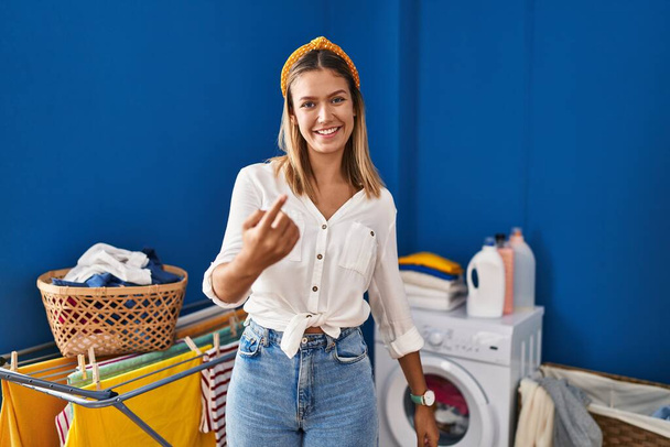 Young blonde woman at laundry room beckoning come here gesture with hand inviting welcoming happy and smiling  - Fotografie, Obrázek