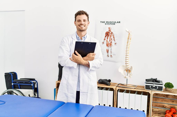 Young hispanic man wearing physiotherapist uniform holding clipboard at clinic - Foto, Bild