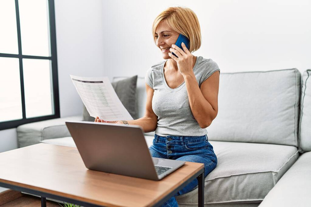 Middle age blonde woman talking on the smartphone using laptop at home - Foto, Bild
