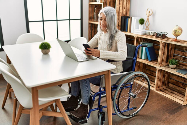Mulher de cabelos grisalhos de meia idade usando smartphone sentado em cadeira de rodas em casa - Foto, Imagem