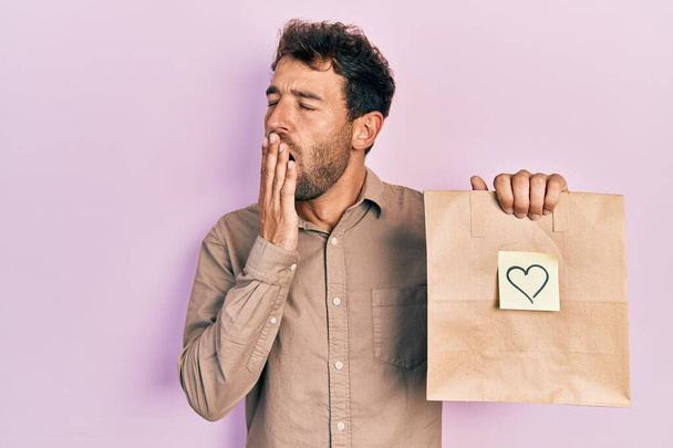 Handsome man with beard holding delivery paper bag with heart reminder bored yawning tired covering mouth with hand. restless and sleepiness.  - Photo, Image