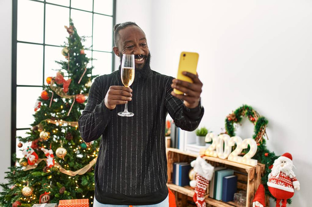 Молодой африканский американец делает селфи на смартфоне, тост с шампанским, стоящим у рождественской елки дома - Фото, изображение