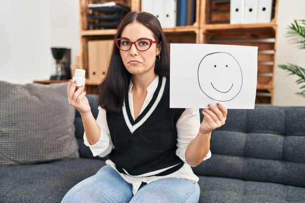 Young hispanic woman working on depression holding happy emotion on paper and pills skeptic and nervous, frowning upset because of problem. negative person.  - Photo, Image