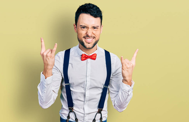 Hispanic man with beard wearing hipster look with bow tie and suspenders shouting with crazy expression doing rock symbol with hands up. music star. heavy concept.  - Photo, Image
