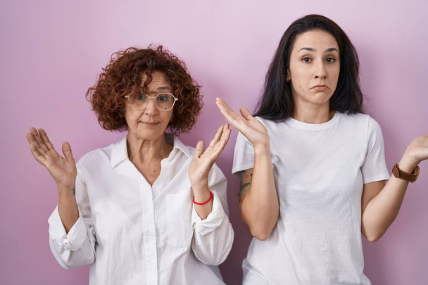 Hispanic mother and daughter wearing casual white t shirt over pink background clueless and confused expression with arms and hands raised. doubt concept.  - Foto, Bild
