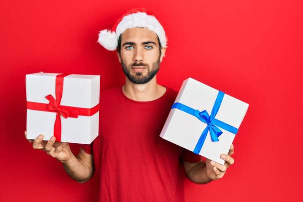 A fiatal spanyol férfi karácsonyi kalapot visel, és ajándékokat tart lazán, komoly arckifejezéssel. egyszerű és természetes nézi a kamerát.  - Fotó, kép