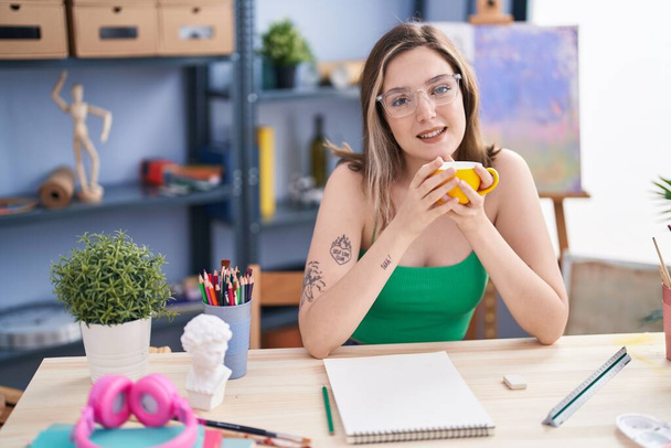 Νεαρή γυναίκα καλλιτέχνης ζωγραφίζει στο σημειωματάριο πίνοντας καφέ στο στούντιο τέχνης - Φωτογραφία, εικόνα