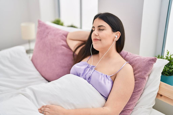 Young hispanic woman listening to music sitting on bed at bedroom - Photo, Image