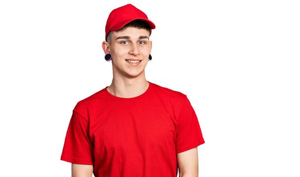 Jeune garçon caucasien avec dilatation des oreilles portant l'uniforme de livraison et le chapeau regardant positif et heureux debout et souriant avec un sourire confiant montrant des dents  - Photo, image