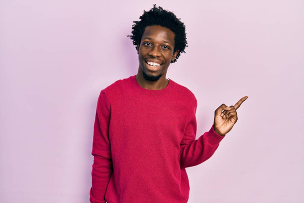 Jeune homme afro-américain portant des vêtements décontractés avec un grand sourire sur le visage, pointant du doigt vers le côté en regardant la caméra.  - Photo, image