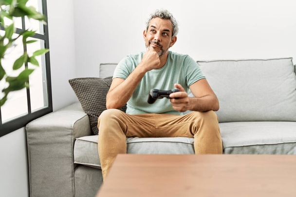 Moyen Age homme hispanique jouer à un jeu vidéo assis sur le canapé visage sérieux penser à la question avec la main sur le menton, réfléchi sur l'idée déroutante  - Photo, image