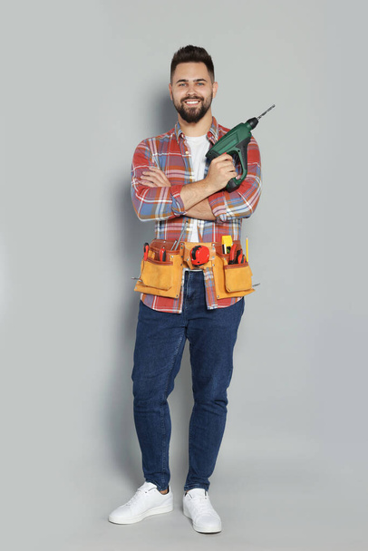 Young worker with power drill and tool belt on grey background - Foto, Bild