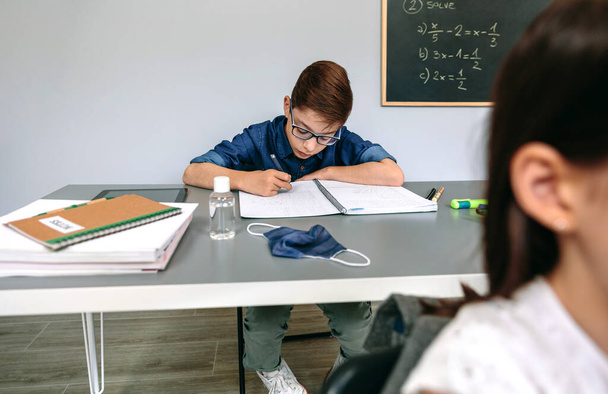 Fiú ír a jegyzetfüzetébe az iskolában maszkkal és kézfertőtlenítővel az asztalon. - Fotó, kép