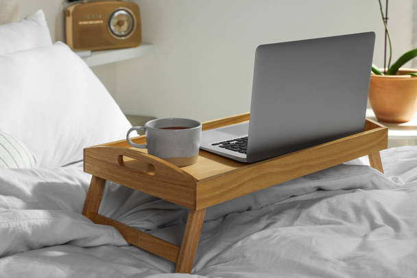 Wooden tray with modern laptop and cup of aromatic tea on bed indoors - Valokuva, kuva