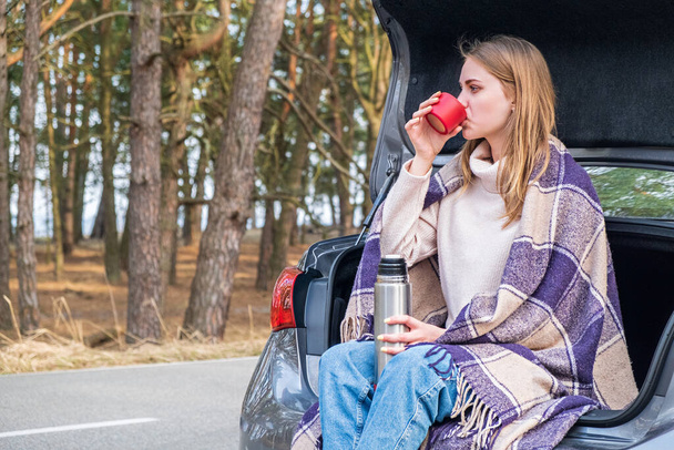 happy young girl sitting in a car trunk drinks hot tea from a thermos cup against the backdrop of a spring or autumn forest. Mental health. Time for yourself, contemplation, meditation. - Photo, Image