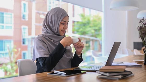 Νεαρή επιχειρηματίας hijab κρατώντας φλιτζάνι καφέ και διαβάζοντας e-mail ή σε απευθείας σύνδεση ειδήσεις στον υπολογιστή laptop. - Φωτογραφία, εικόνα
