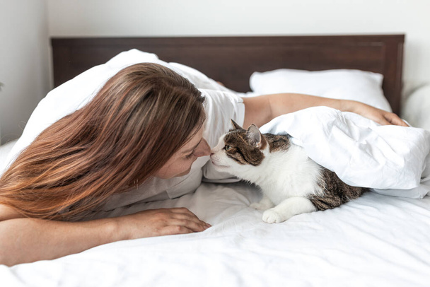 Happy young woman with cat in bed at home. In cold weather, the pet warms up under a blanket. Pet friendly and grooming concept. Stray kitten sleep on bed. Cozy home background - Foto, afbeelding