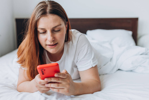 Schöne junge Frau liest SMS auf ihrem Handy im Bett. Soziale Netzwerke checken, SMS verschicken. Das Mädchen trägt ein T-Shirt. - Foto, Bild