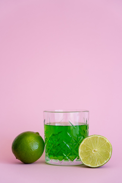 green alcohol drink in faceted glass near fresh limes on pink background - Zdjęcie, obraz