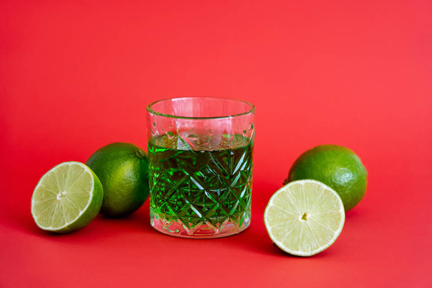 green alcohol drink in faceted glass near sour limes on red - Foto, imagen