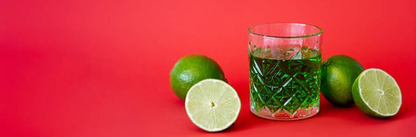green alcohol drink in faceted glass near sour limes on red, banner - 写真・画像