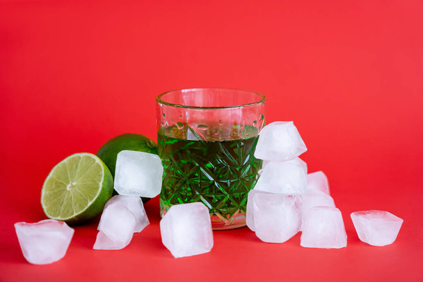 замороженные кубики льда возле стекла со спиртом зеленый мохито и лаймы на красном - Фото, изображение