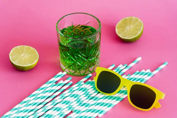 napszemüveg és egy üveg zöld alkoholtartalmú ital csíkos papírszívószál és -mész közelében, rózsaszínen  - Fotó, kép