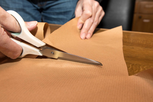 A closeup of a person's hands cutting a paper with scissors for a gift package on a wooden table - Foto, afbeelding