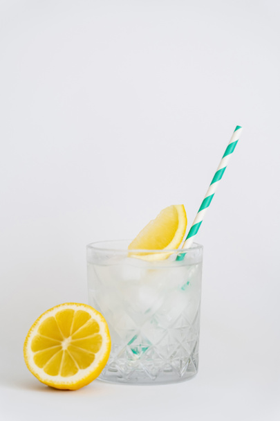 faceted cold glass with ice cubes, paper straw and sliced lemons on white - Фото, изображение