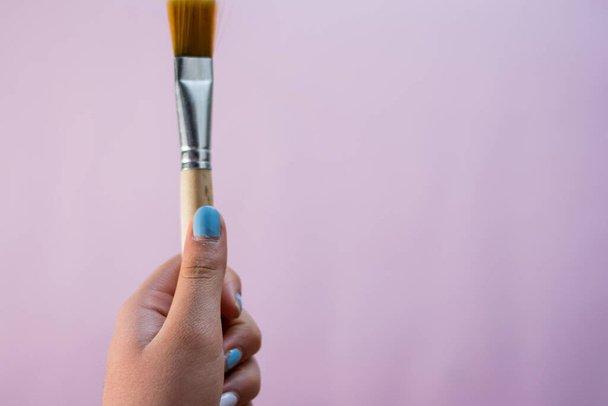 A closeup of a female hand holding a painting brush on a pink background with copyspace - Foto, afbeelding
