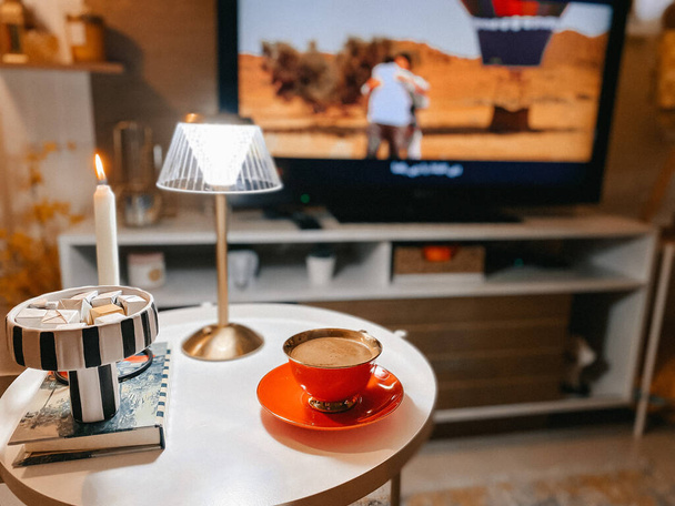 A closeup of beautifully arranged a cup of coffee on a table decorated with a lamp against a TV - Fotó, kép