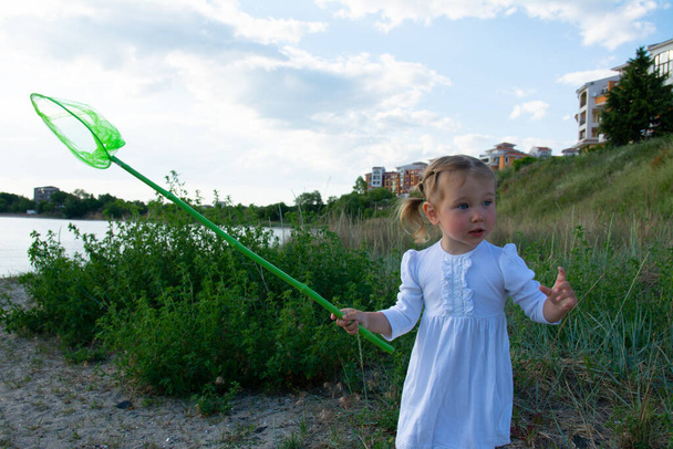 Piękna dziewczynka w białej sukience z zieloną siatką w rękach na plaży i na tle zielonej trawy i błękitnego nieba. Piękna dziewczynka z siatką. - Zdjęcie, obraz