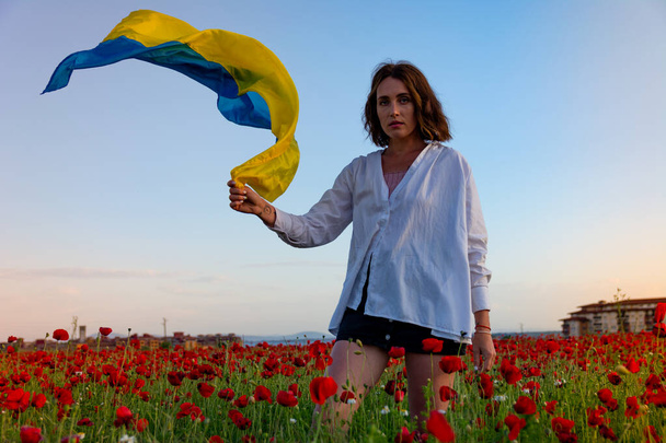 Piękna młoda dziewczyna w białej koszuli i szortach pozuje na polu maku z flagą Ukrainy o zachodzie słońca. - Zdjęcie, obraz