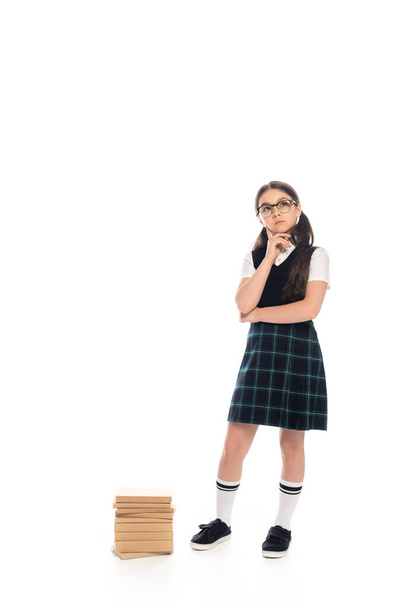Pensive schoolkid in skirt and eyeglasses standing near books on white background - Photo, Image