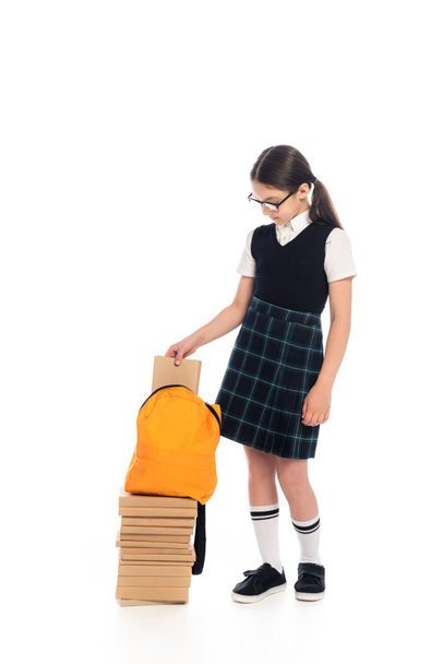 Full length of preteen schoolgirl putting book in backpack on white background - Foto, immagini