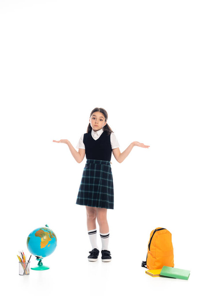 Confused schoolchild showing shrug gesture near globe and books on white background - Фото, зображення
